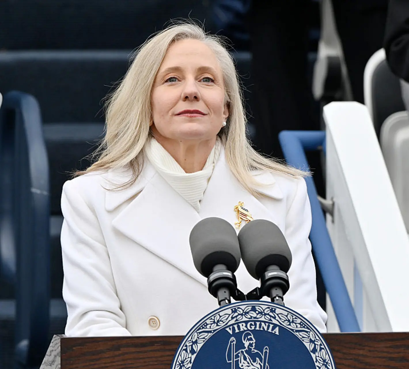 A photo of Spanberger at a podium dressed in a fancy stark white suit and an imperious smirk like some kind of Hunger Games villain.